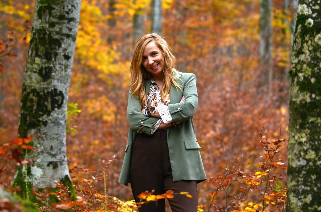 Ženska z dolgimi lasmi, oblečena v zeleno jakno in rjav spodnji del, se nasmejana postavlja v gozdnem okolju s pisano jesensko vegetacijo.